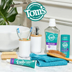 Bathroom scene with Tom’s of Maine toothpaste tube in foreground and other Tom’s products near a sink and towel