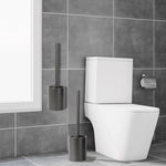 Bathroom scene showing a gray toilet brush and holder placed by a toilet and an identical holder mounted on tiled wall