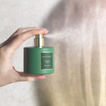 Hand holding and spraying a small green BEAUTYSPACE Winter Pine room spray with mist visible against a neutral background