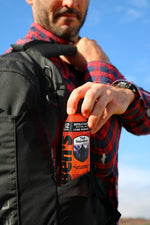 Person placing an orange can of Ben's Tick Repellent into a backpack pocket while wearing a red plaid shirt outdoors.