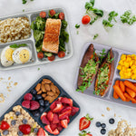 Overhead lifestyle view of several Bentgo Prep three-compartment containers filled with various meals and scattered herbs and fruit