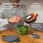 Lifestyle image of person assembling Bentgo compartment tray over a glass salad bowl on a wooden table with lid and sauce cup visible