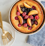 Baked cake with peach slices, blackberries and raspberries served in a round aluminum pan next to forks and a napkin