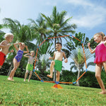 Group of children playing and jumping through an orange and blue octagon sprinkler set up on a sunny lawn.