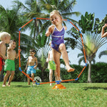 Young girl jumping through an orange and blue octagon sprinkler while other children cheer on a grassy yard.