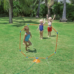 Octagonal orange and blue sprinkler standing on grass with three children in swimsuits playing behind it.