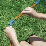 Close-up of hands connecting orange tubing to a blue elbow joint on a plastic sprinkler frame over grass.