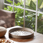 Lifestyle image of a gray-brown Better Homes & Gardens ceramic planter saucer placed on a table near a window with plants in background.