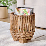 Woven water hyacinth footed planter used as a magazine holder beside a sofa on a light rug