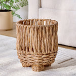 Close view of a woven water hyacinth planter showing braided texture and short rounded feet on a rug
