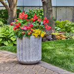 Outdoor scene showing the gray fluted gypsum planter filled with colorful flowering annuals on a paved patio