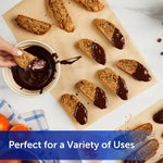 Biscotti pieces on unbleached parchment paper with a bowl of melted chocolate and a hand dipping a cookie