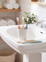 Pastel Bizzu training toothbrushes displayed in a white cup on a bathroom sink with towels and plant in background