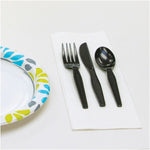 White napkin with a black plastic fork, knife and spoon beside a patterned paper plate on a white table.