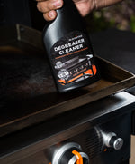 Hand holding a Blackstone Degreaser Cleaner bottle angled over a griddle, label clearly visible