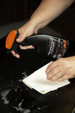 Person spraying Blackstone Degreaser Cleaner and wiping a griddle surface with a paper towel