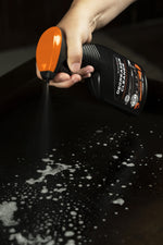 Hand holding a Blackstone Degreaser Cleaner bottle spraying foam onto a dark griddle surface