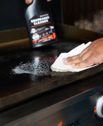 Close view of a hand wiping a foamy area on a griddle with a blurred Blackstone Degreaser Cleaner bottle in background
