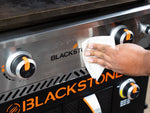 Hand wiping the stainless steel front panel of a Blackstone griddle near control knobs with a paper towel