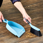 Hands using a blue dustpan and matching small broom to sweep up dry crumbs from a wood floor