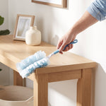 Hand holding a blue-handled duster cleaning the edge of a wooden table in a bright home setting