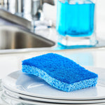 Blue non-scratch scrub sponge resting on a stack of white plates with a blurred kitchen sink and soap dispenser in the background.