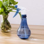 Blue pear-shaped refillable spray bottle on a wooden surface next to a small glass vase of green plants.