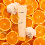 Boka Kids Orange Cream toothpaste tube staged on a background of sliced oranges with scoops of orange-cream ice cream for flavor context.