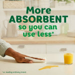 Hand wiping a kitchen counter with a paper towel next to a spilled yellow liquid and a glass of orange juice