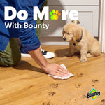 Person wiping muddy paw prints from a hardwood floor with a Bounty sheet while a puppy watches nearby