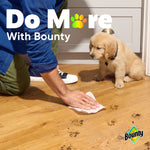 Hand using a Bounty paper towel to wipe muddy paw prints from a hardwood floor while a puppy watches
