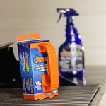 Boxed orange-handled Citrusafe grill scrubber in focus with a blurred Citrusafe spray bottle in the background.