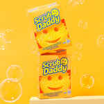 Two boxed Scrub Daddy smile-face sponges stacked on a bright yellow background with soap bubbles