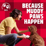 Lifestyle-style image showing a woman wiping a curly dog’s paw against a red plaid background with text about muddy paws.