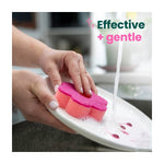 Hand holding pink flower-shaped scrub sponge rinsing a sudsy plate with caption ‘Effective + gentle’