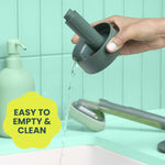 Hand pouring water from the round base stand while a Brite dishwand and scrubber lie on the counter in a tiled sink scene