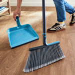 Lifestyle photo of a turquoise broom and matching dustpan on a wood floor with a person reaching for the dustpan
