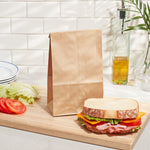Single brown kraft paper lunch bag standing on a cutting board beside a sandwich and sliced tomato in a kitchen setting.