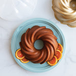 Top-down styled bundt cake on a teal base with orange slices, a gold bundt pan and clear dome lid nearby