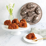 Staged scene with a bunny-cavity cake pan propped upright and baked bunny cakes on a white pedestal