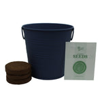 Navy metal pail with two compressed soil discs and a Buzzy seeds packet arranged beside it on white