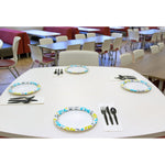 Cafeteria-style table set with paper plates and black plastic utensils placed at multiple settings in a dining area.