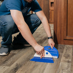 Person using a Camco blue dust pan and whisk broom to sweep on a wood-look floor