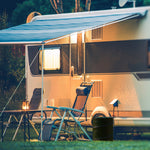 Campground scene with a camper, chairs and a dark green collapsible trash can placed beside the seating area