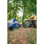 Campsite scene with a green Stansport collapsible trash can in the foreground and people gathered near tents and a camper van.