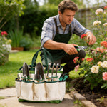 Lifestyle photo of a canvas tool bag filled with metal hand tools on a path while a gardener prunes nearby flowers