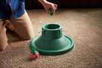Kneeling person placing a small green cap over a tree stand with a Miracle-Gro bottle resting beside it on the carpet.