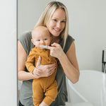 Caregiver brushing a baby's mouth with a gray silicone finger toothbrush while looking into a mirror in a bathroom