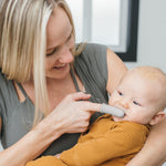 Lifestyle image of a caregiver using a silicone finger toothbrush on a baby held in their arms in a softly lit room.