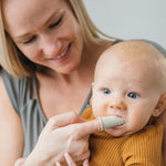 Adult placing a silicone finger toothbrush into a baby’s mouth while smiling, close-up lifestyle shot showing baby and caregiver.